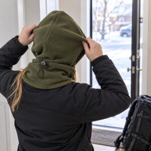 A woman in a black jacket pulls up an olive green fleece hood with a cord toggle at the neck, standing by a window in winter.